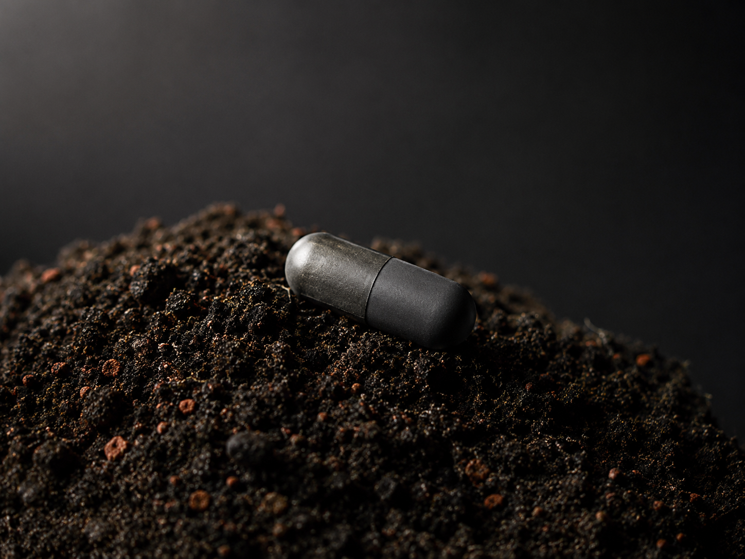 A single dark matte capsule resting on a mound of dark soil flecked with terracotta-red grains — visualizing the bridge from a 1971 soil microbe to a daily oral postbiotic. Illustrative placeholder image, to be replaced with a product photograph.